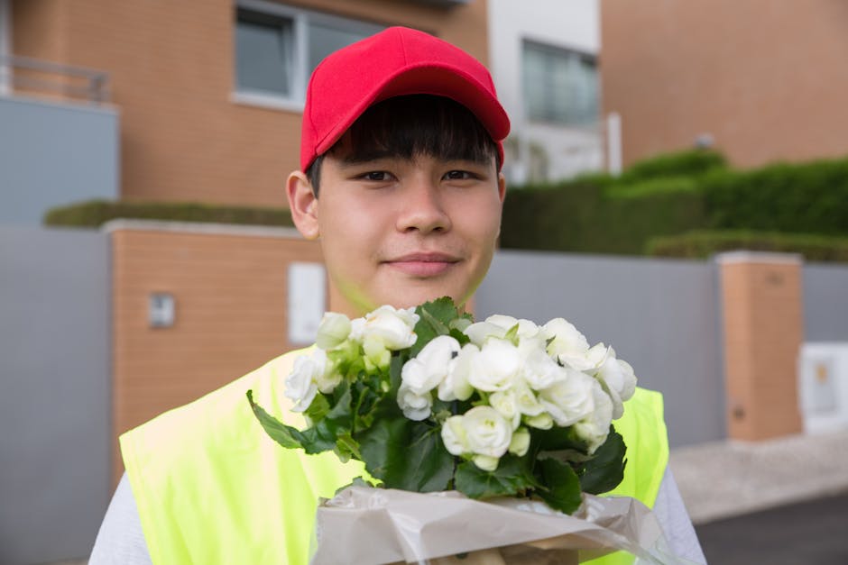 A smiling young delivery person wearing a red cap and high-visibility yellow vest is holding a bouquet of white roses wrapped in white paper in front of a modern residential building. The bouquet feat