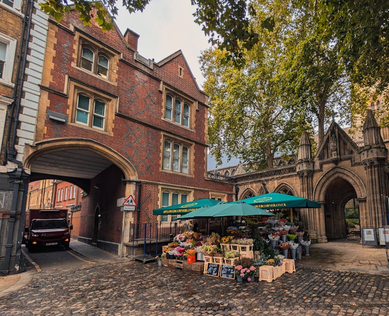 A flower stall set up outside a historic red-brick building with arched entryways, displaying a wide variety of fresh flowers including pink roses, white lilies, yellow tulips, and mixed seasonal bloo