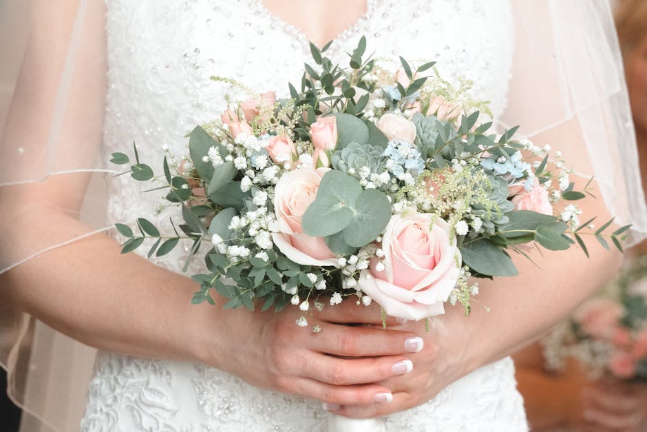 A bride holding a bridal bouquet composed of soft pink roses, baby's breath, and eucalyptus leaves, arranged in a rounded style, with the roses prominently displayed at the center. The bouquet feature